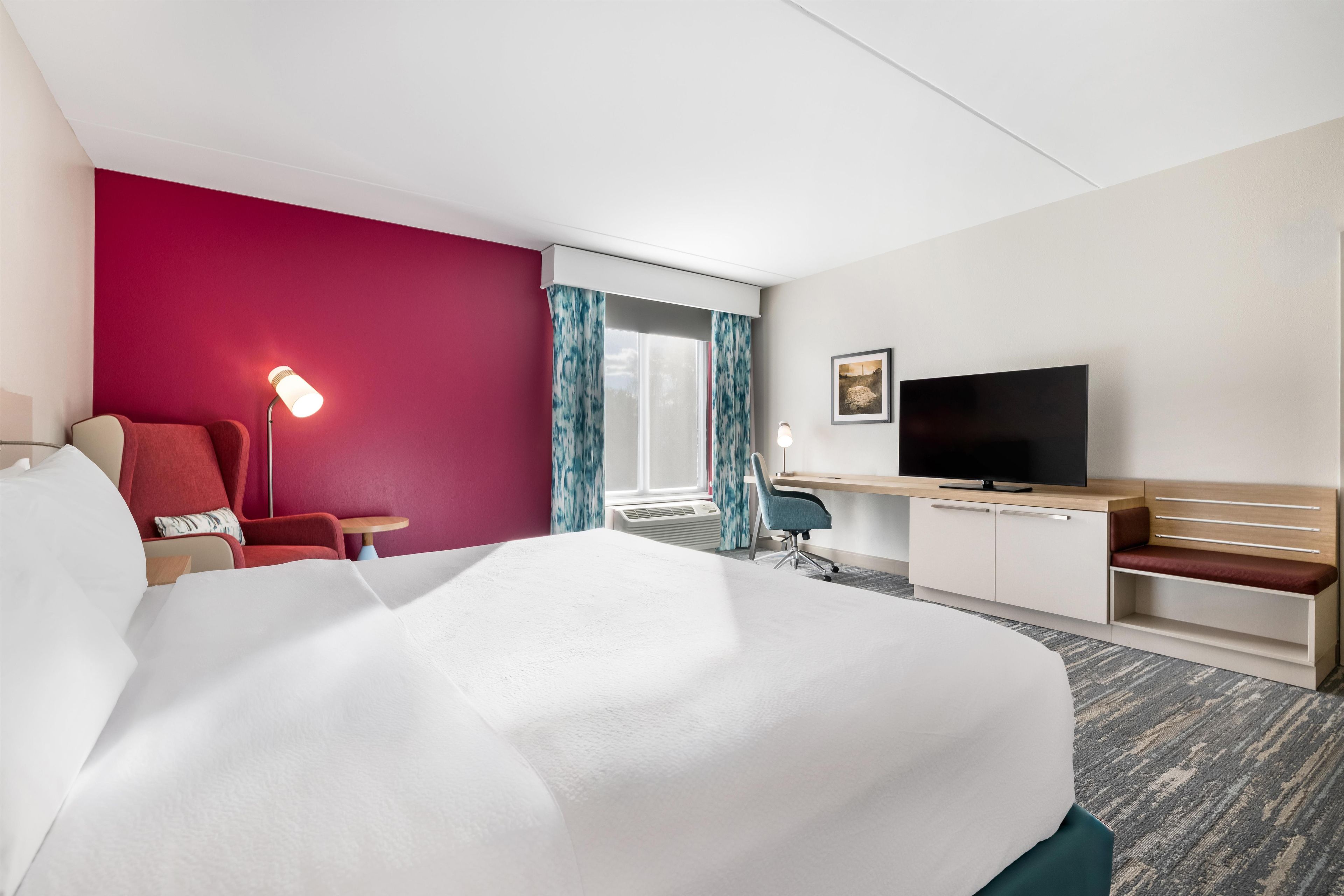 Ein Hotelzimmer mit einem großen Bett, einem Schreibtisch mit Flachbildfernseher, einem Sessel, einer Schreibtischlampe und einem Fenster mit Vorhängen.