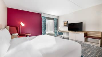 Ein Hotelzimmer mit einem großen Bett, einem Schreibtisch mit Flachbildfernseher, einem Sessel, einer Schreibtischlampe und einem Fenster mit Vorhängen.