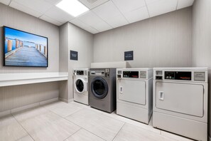 A laundry facility with a large wall-mounted TV, multiple washing machines, and dryers. - Hilton Garden Inn Mystic/Groton (Groton)
