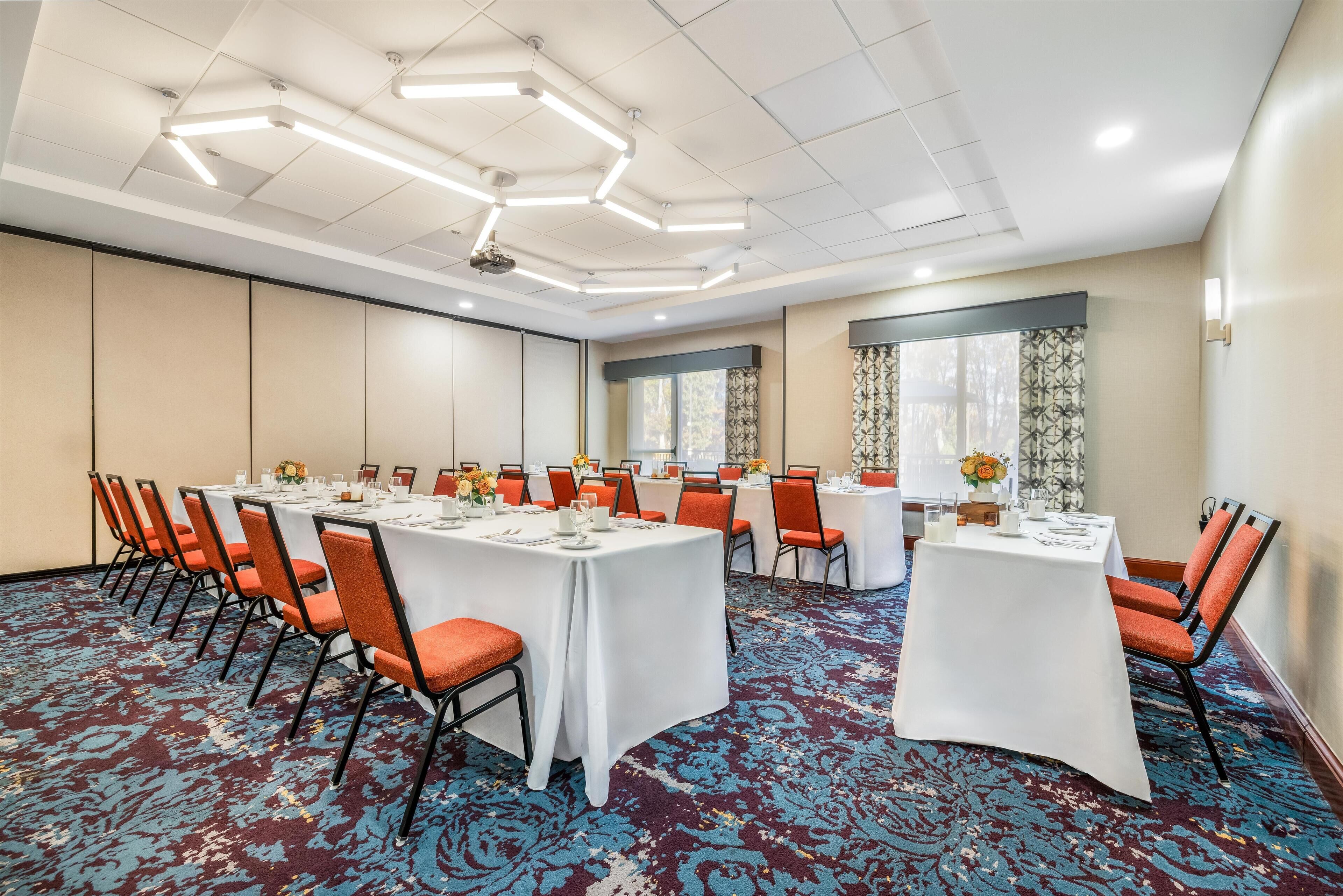 Una sala de conferencias con una larga mesa dispuesta para una reunión, rodeada de sillas anaranjadas, un suelo con alfombra estampada e iluminación empotrada en el techo.