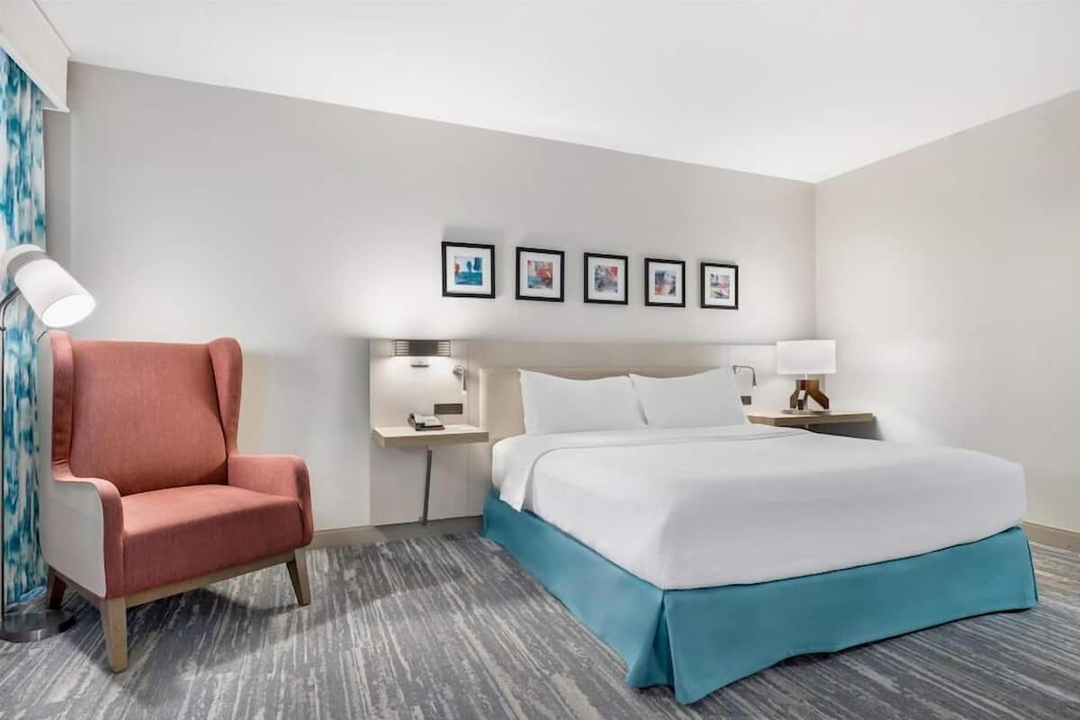 A hotel room with a bed, a chair, a nightstand, a lamp, and framed pictures on the wall.