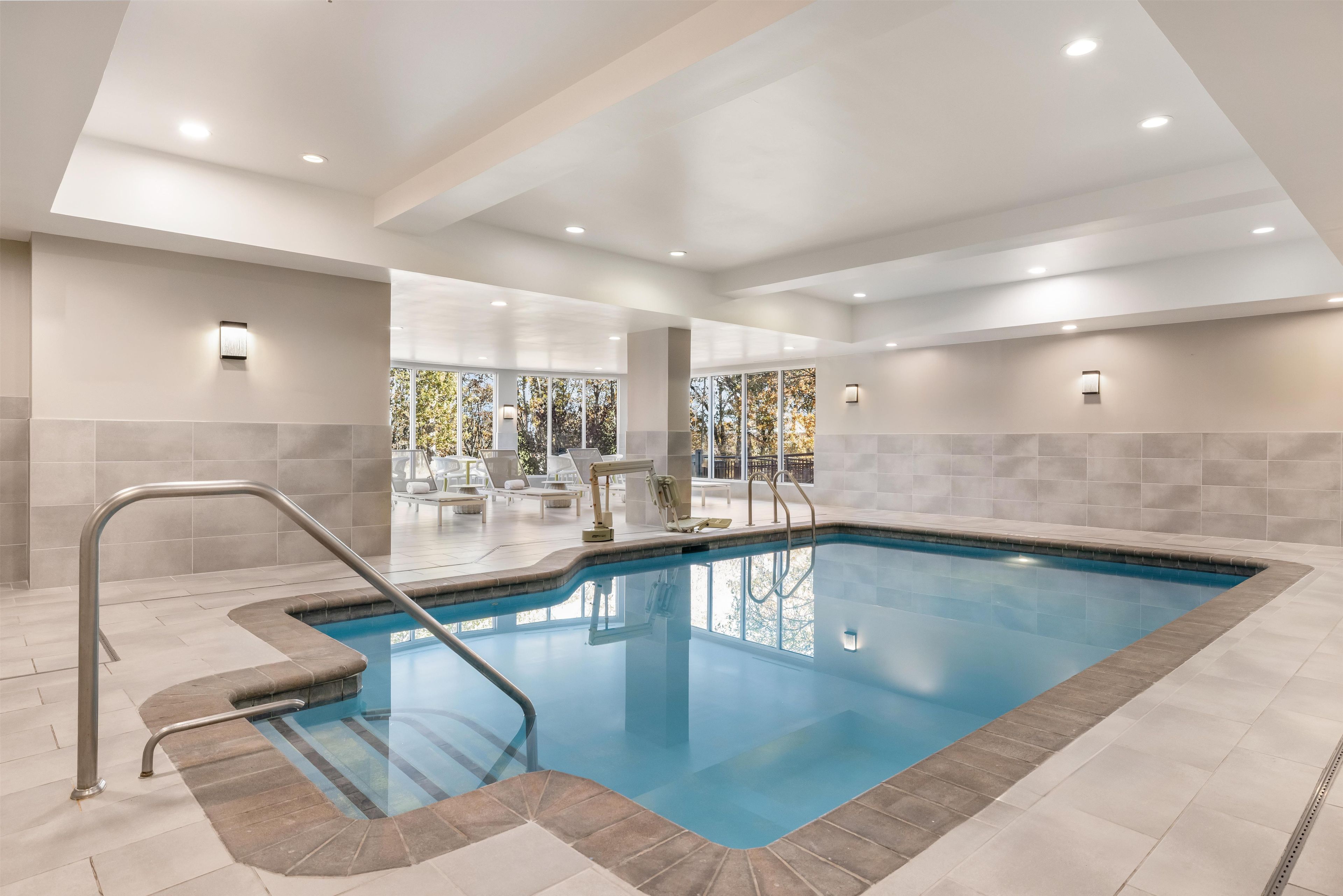 Piscina interior moderna com água azul cristalina, rodeada por paredes revestidas e uma área ensolarada com espreguiçadeiras.