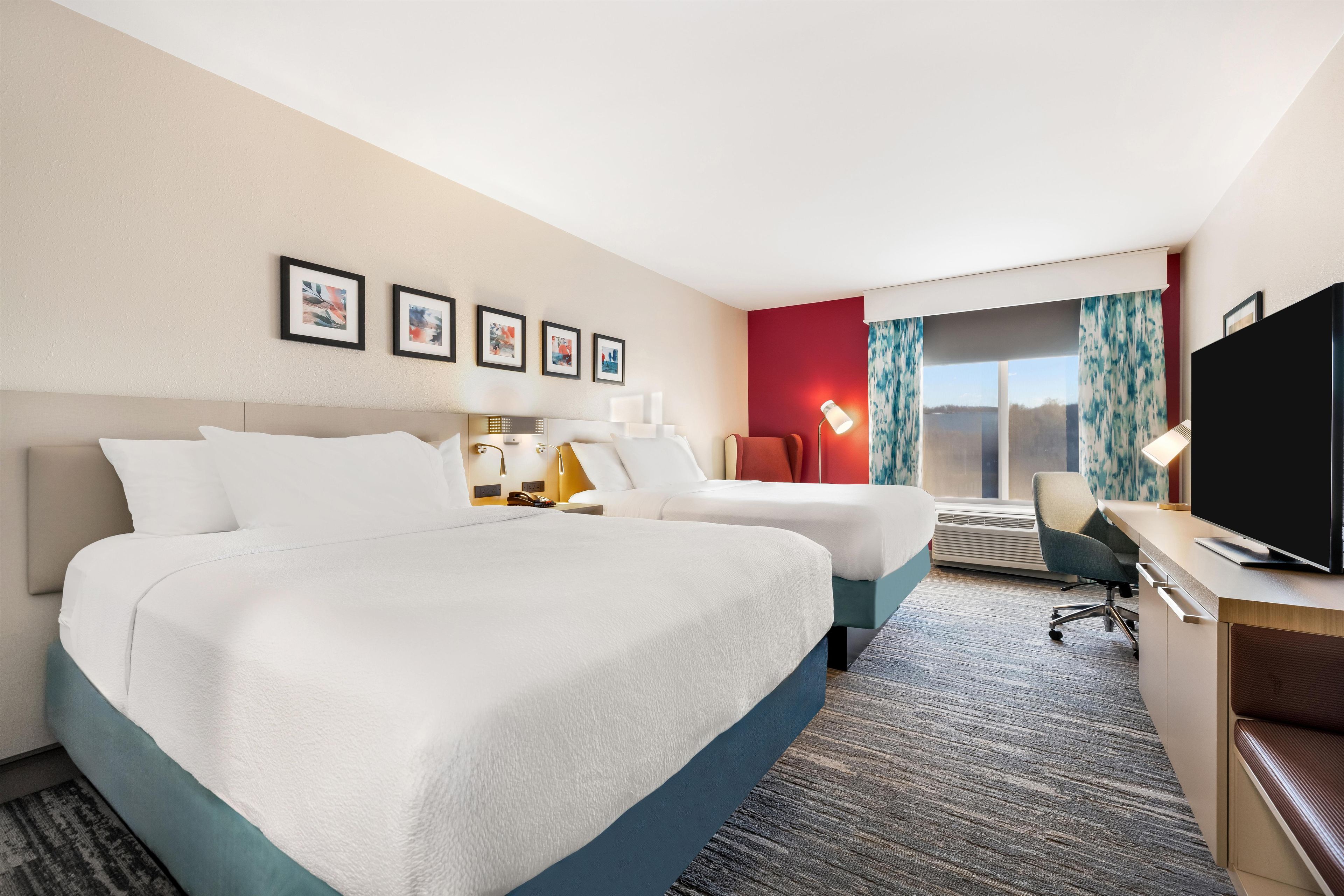 A hotel room with two beds, a desk with a television, a window with curtains, and wall-mounted artwork.
