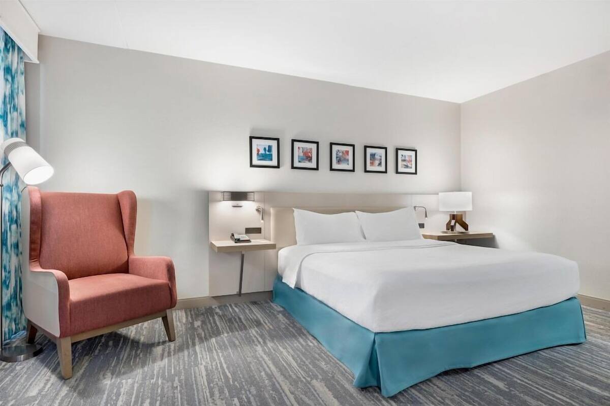 A hotel room with a bed, a chair, a nightstand, a lamp, and framed pictures on the wall.