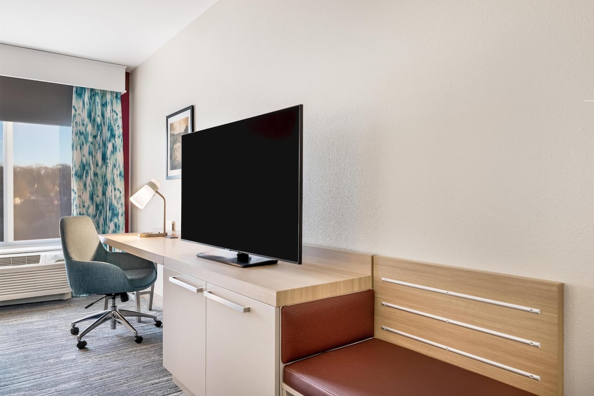 A modern hotel room with a wooden desk, a large flat-screen TV, a comfortable chair, a bedside table, and a window with a view.