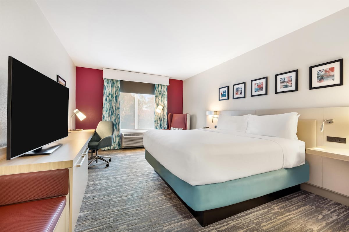 A hotel room with a large bed, a desk with a flat-screen TV, a chair, a desk lamp, and framed pictures on the wall.