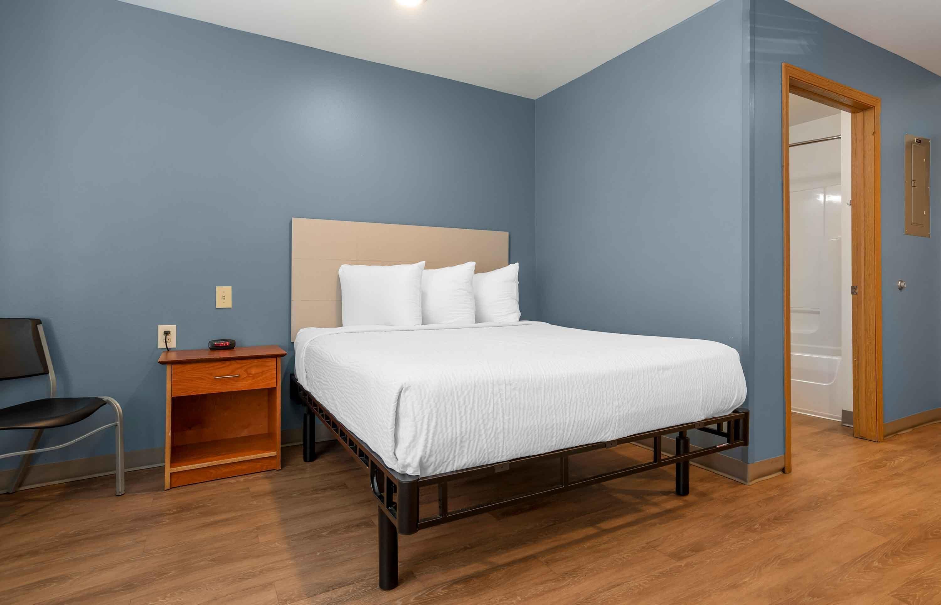 Um quarto com uma cama, uma mesinha de cabeceira, uma cadeira e uma porta de madeira.