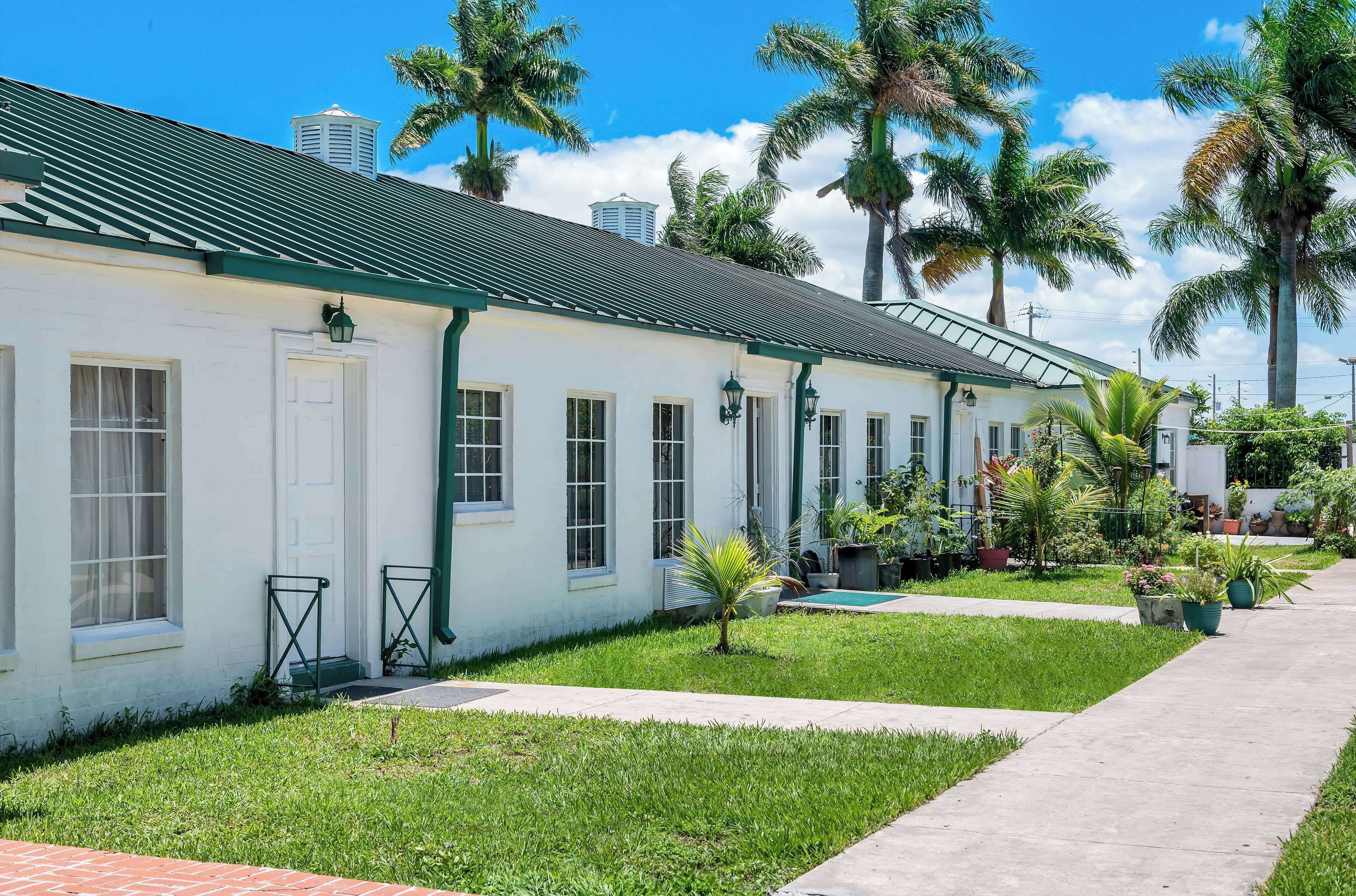 A row of white buildings with green trim and a tiled roof, surrounded by palm trees and a well-maintained lawn.