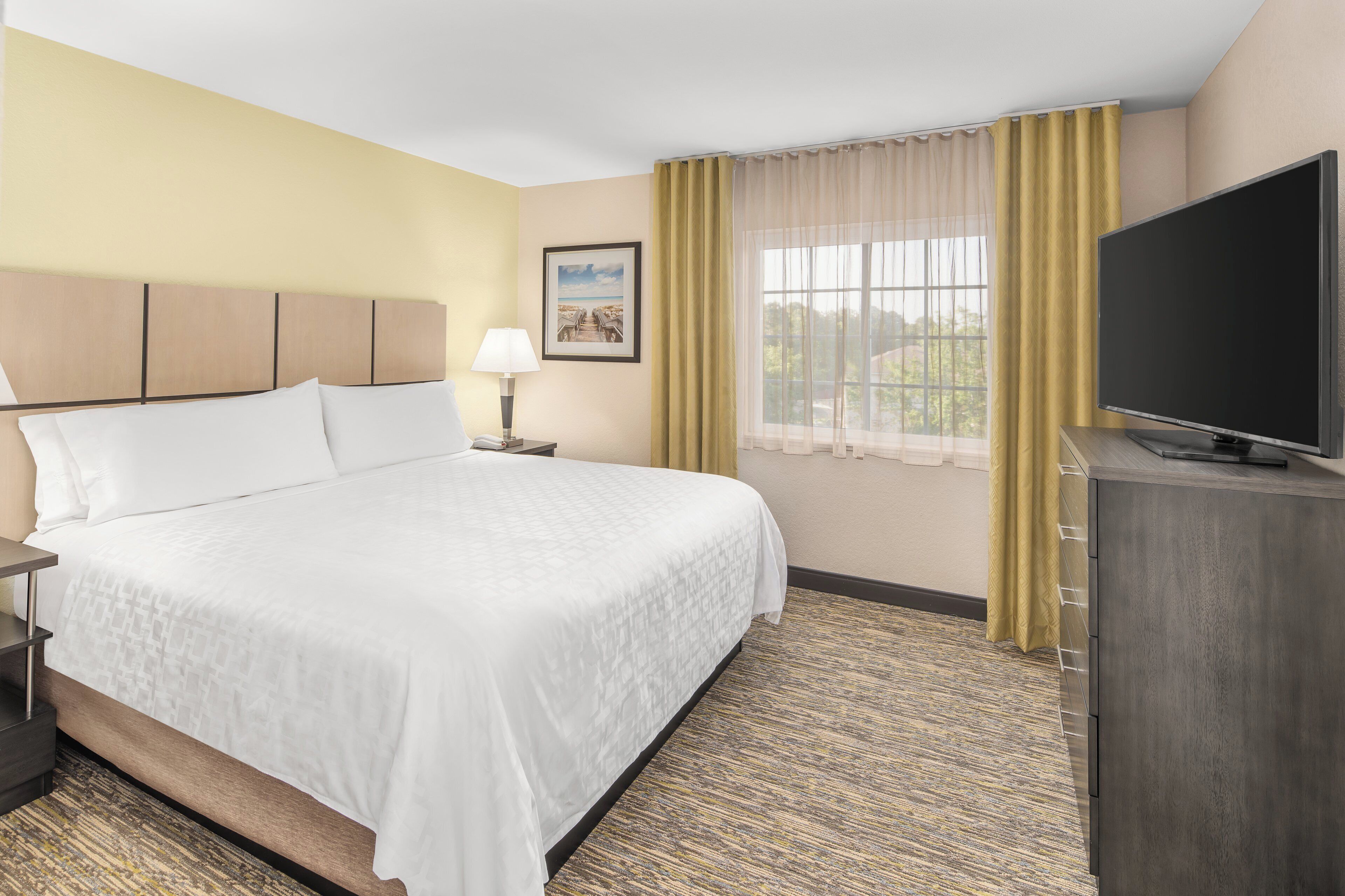 A hotel room with a large bed, a flat-screen TV, a bedside table, a lamp, and a window with curtains.