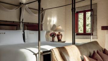 Una cama con dosel con ropa de cama blanca, una mesita de noche con una lámpara y flores, y vistas al follaje a través de una ventana.