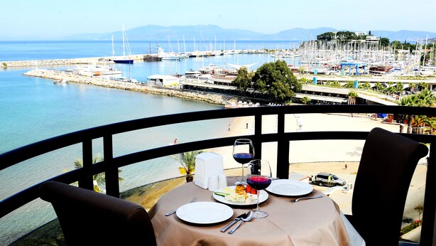 Ein Tisch ist mit Tellern, Besteck und Weingläsern gedeckt, mit Blick auf eine Marina mit Booten und einen Strand.
