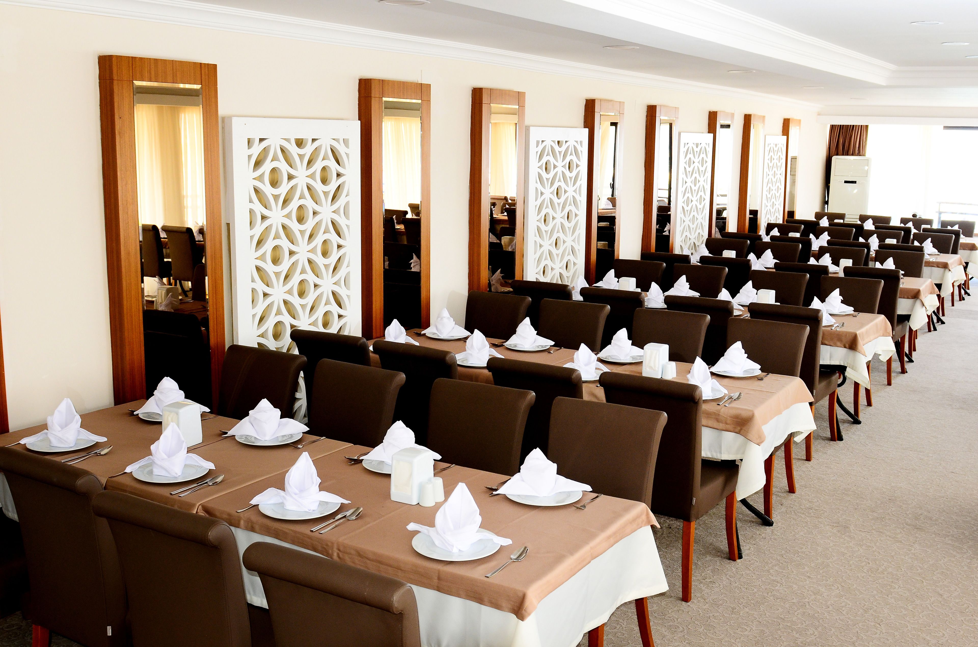 Een restaurantgedeelte van een hotel met meerdere gedekte tafels, elk met een witte tafelkleed, bruine stoelen en netjes opgemaakte servetten en bestek.