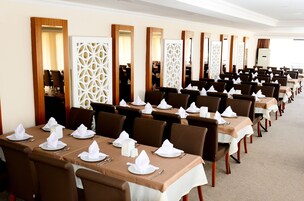 Uma área de refeições de hotel com várias mesas preparadas para o serviço, cada uma com toalha de mesa branca, cadeiras marrons e guardanapos e talheres arrumados.