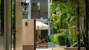 Un étroit sentier mène à un espace aménagé avec un banc et un parasol, le tout entouré d’une végétation luxuriante et d’un bâtiment doté d’un balcon.