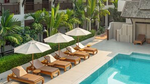 Un espace aménagé au bord de la piscine, avec des chaises longues et des parasols, entouré de verdure et bordé d’un bâtiment doté de balcons.