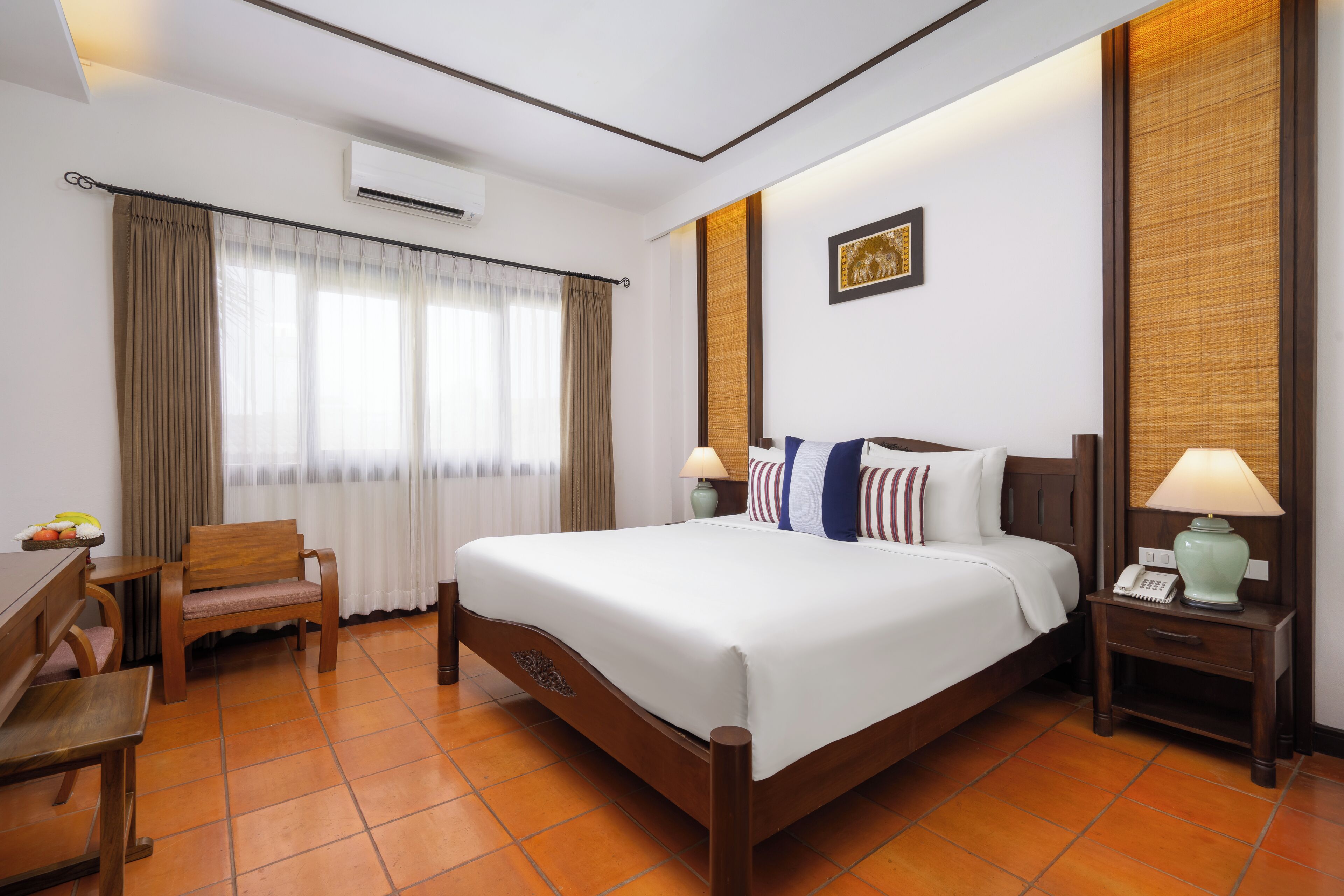 Une chambre d’hôtel avec un lit, une chaise, une table de chevet, une lampe et une fenêtre avec des rideaux.