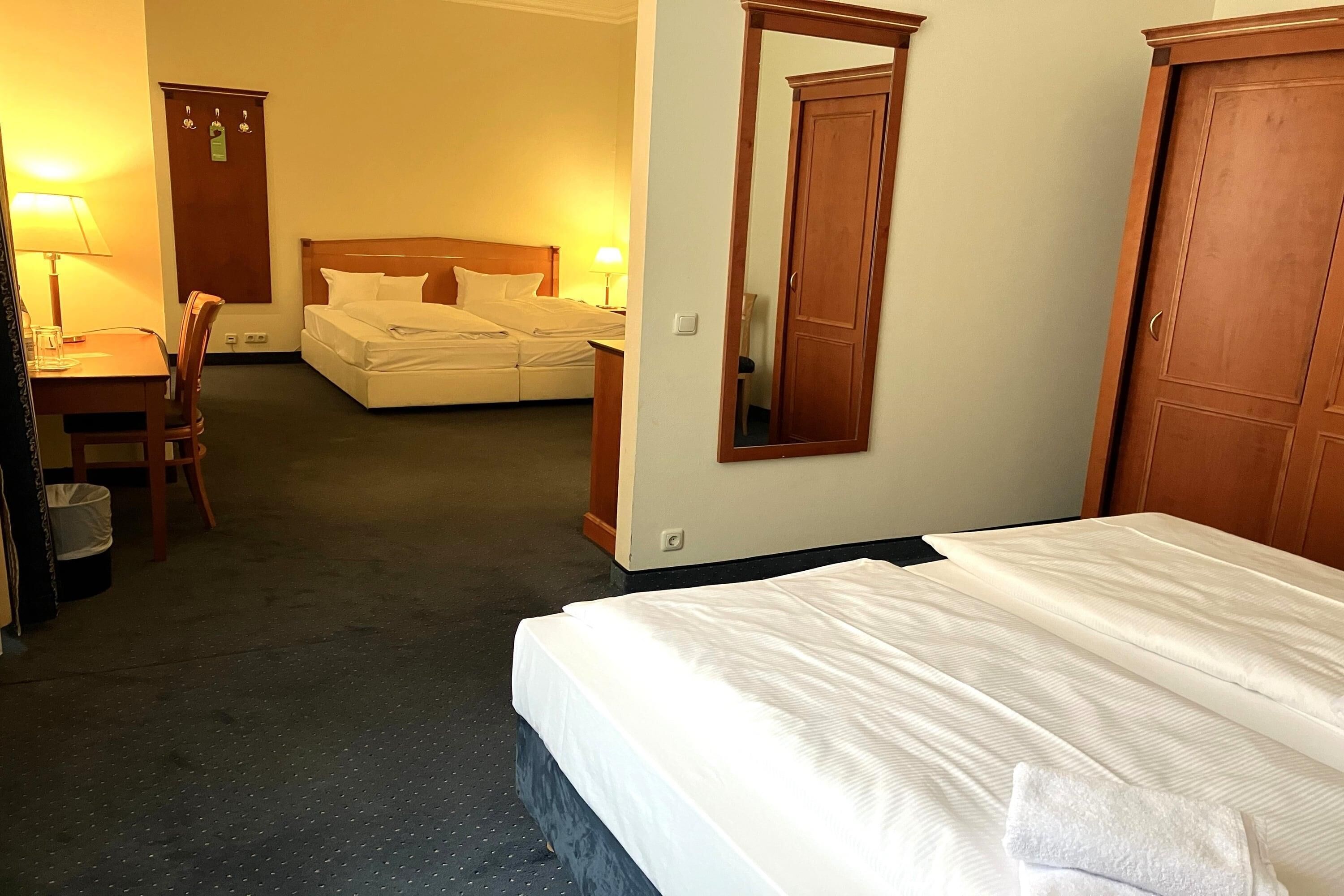 A hotel room with a bed, a desk with a chair, a wardrobe, and a mirror.