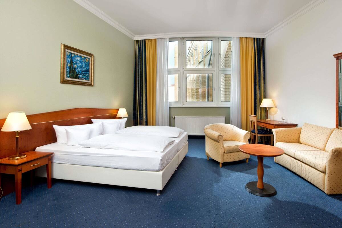 Habitación de hotel con cama, dos mesitas de noche, un sofá, una pequeña mesa redonda y una ventana con cortinas.