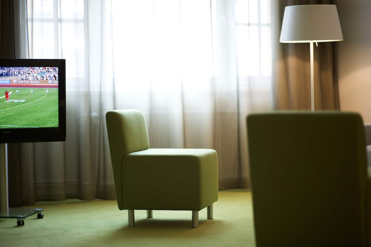 Una habitación de hotel con una televisión mostrando un evento deportivo, un sillón verde y una lámpara.