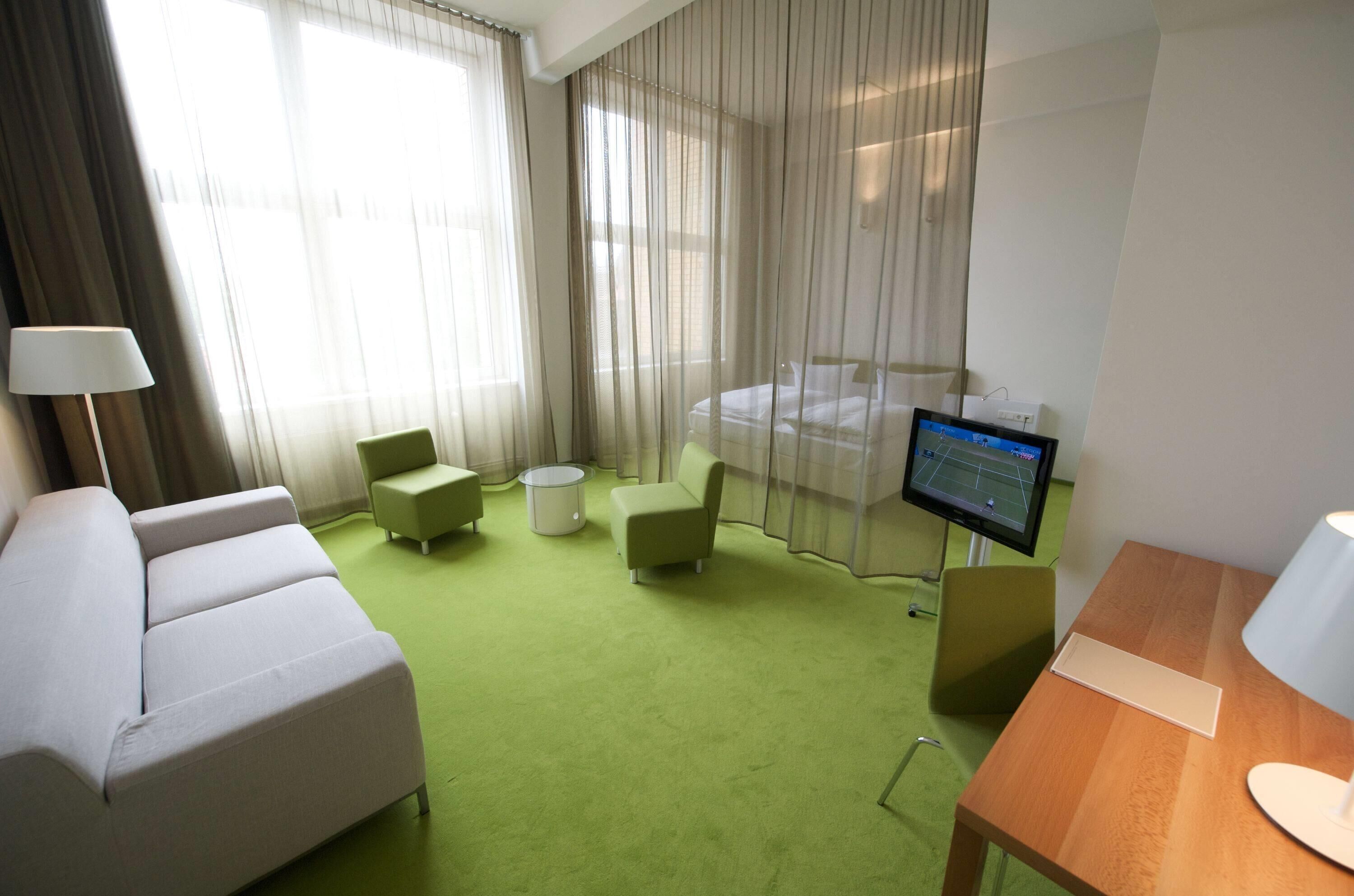 A hotel room with a green carpet, a bed, a TV, a desk, and a chair.