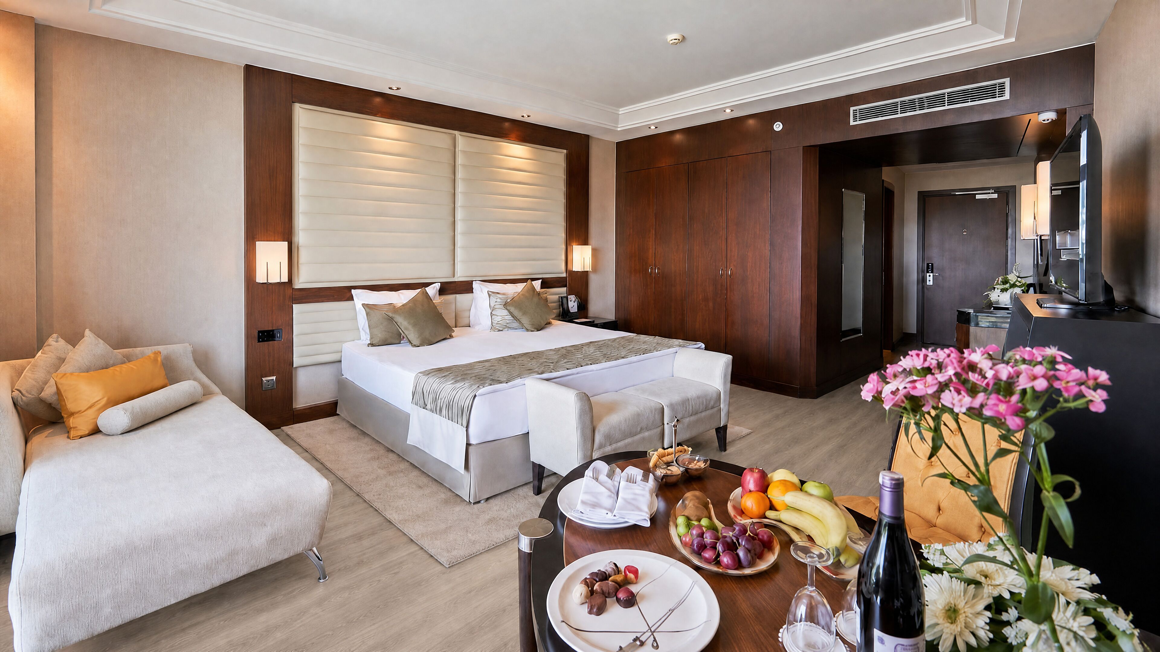 A hotel room with a bed, a sofa, a dining table with fruits and a bottle, and a desk with a computer.