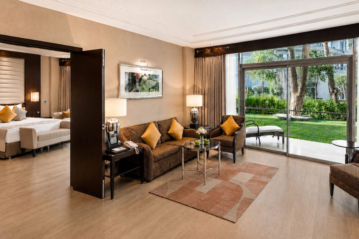 A hotel room with a bed, a sofa, a coffee table, and a TV. There is a large window with curtains and a view of greenery outside.