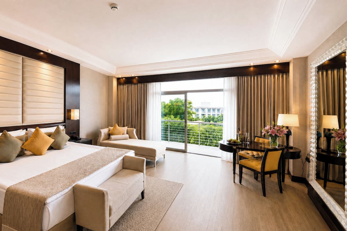 A hotel room with a large bed, a smaller sofa, a dining area with a table and chairs, and a view of greenery outside.
