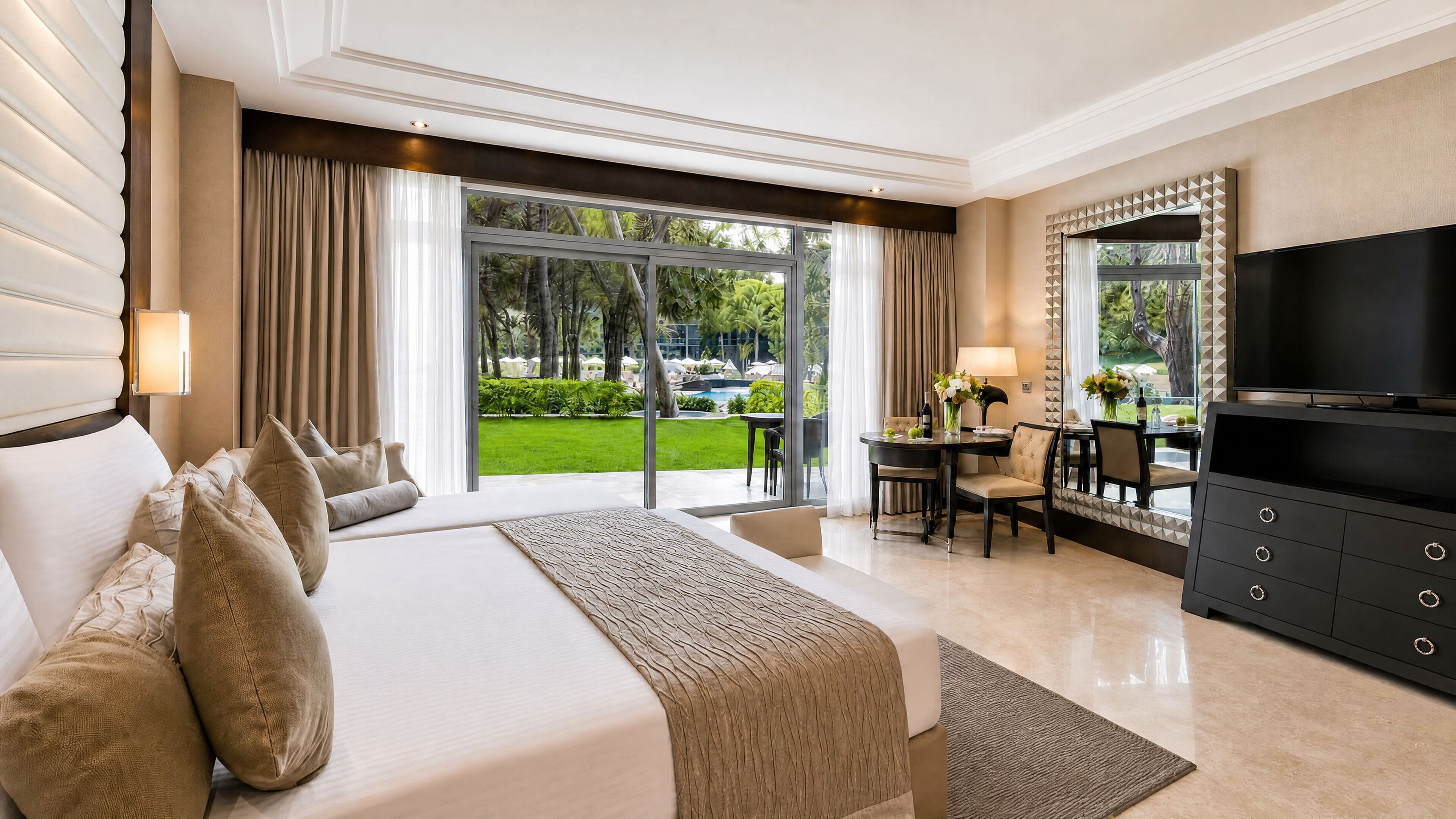 A hotel room with a large bed, a TV, a dining area, and a view of a garden.