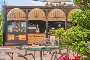 Un espacio al aire libre con mesa de vidrio, un pastel y una taza de café. Al fondo, una cafetería con un letrero que dice "Coffee & Ice Cream".