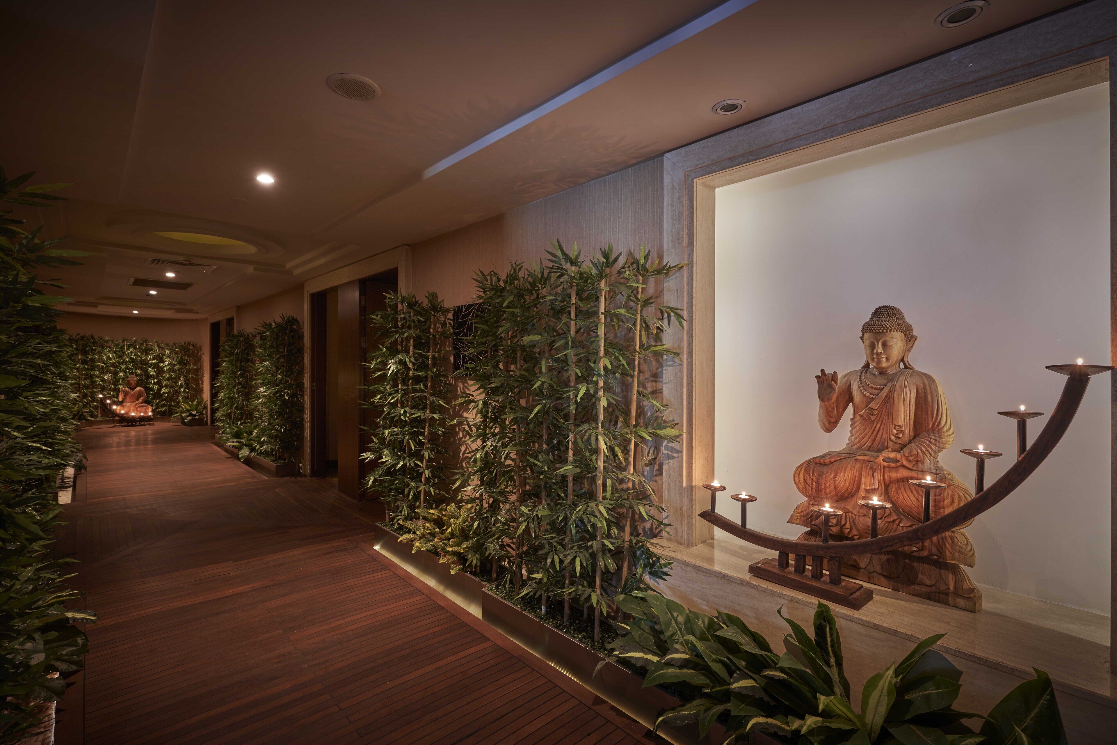 A corridor with a wooden floor, a large Buddha statue on the right, and potted plants along the walls.