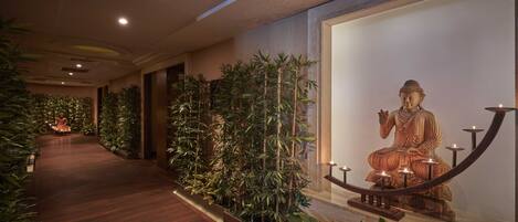 A corridor with a wooden floor, a large Buddha statue on the right, and potted plants along the walls.