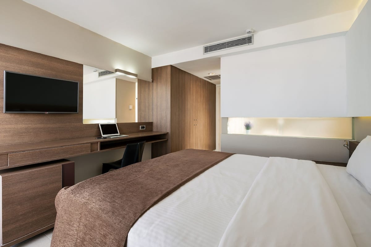 Una habitación de hotel moderna con una cama grande, un escritorio con una computadora portátil, un televisor de pantalla plana en la pared y un espejo amplio.