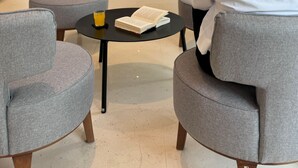 A modern hotel lobby with grey upholstered chairs, a round black table with a book and a glass of orange juice, and a large window offering a view of the outdoors. -  Angela Downtown Hotel  (Rhodes)