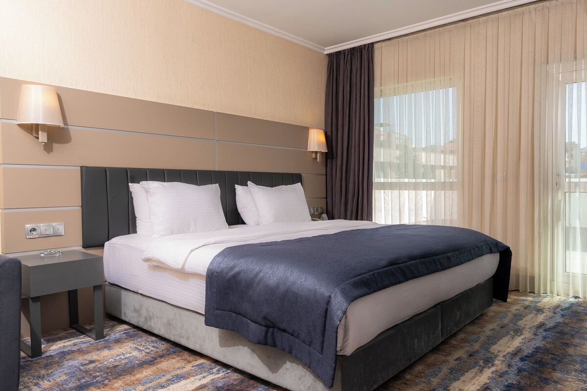 Una habitación de hotel con una cama grande, dos mesitas de noche, una lámpara de noche, una ventana con cortinas y suelo alfombrado.