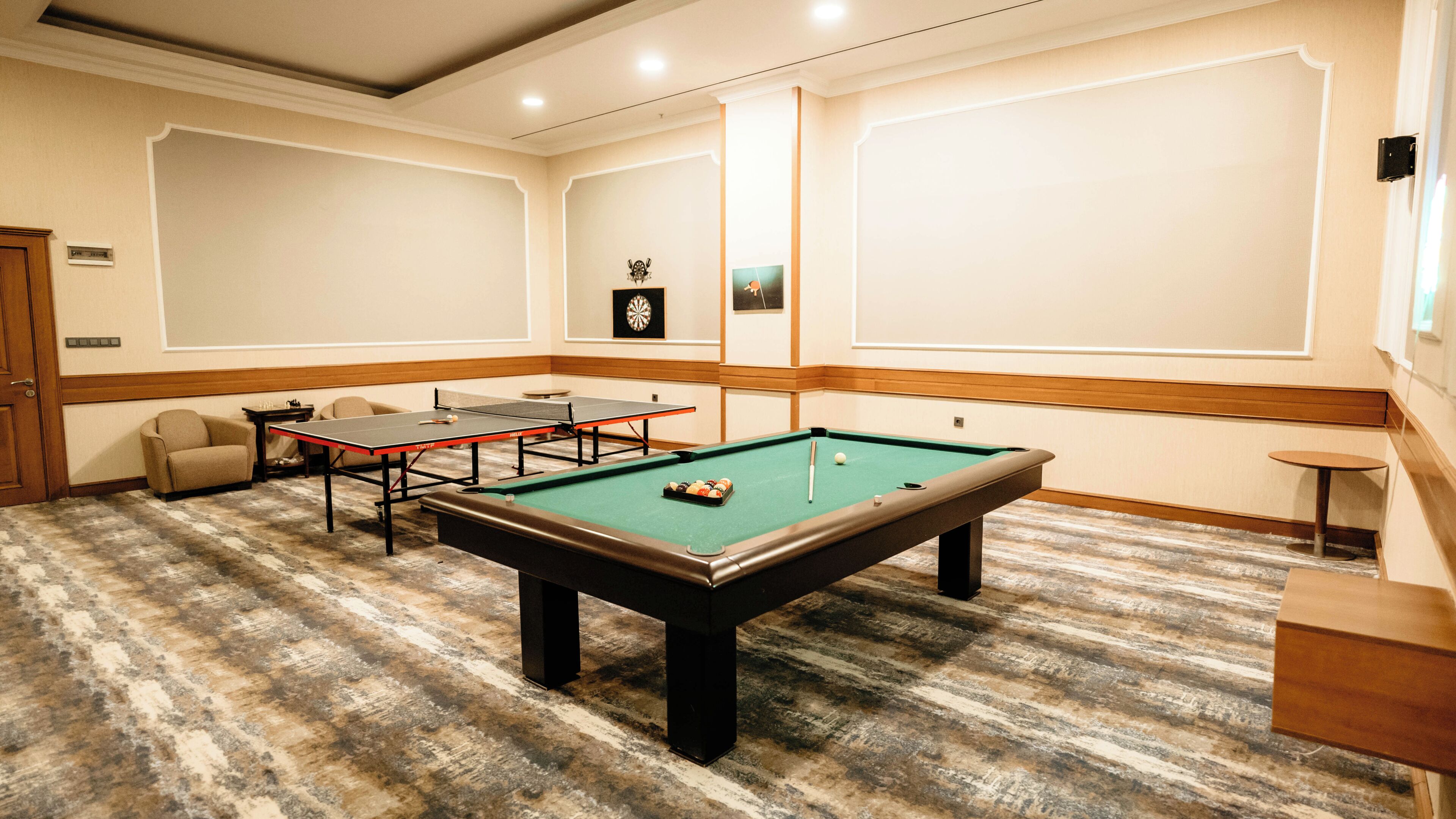 A pool table with a green felt surface, a ping pong table, and a billiards table in a room with patterned carpet flooring and wooden paneling.
