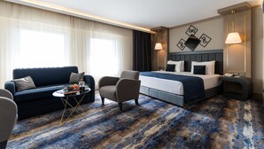 Una habitación de hotel con una cama, un sofá, una silla, una mesita con frutero y una lámpara.