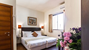 A hotel room with a bed, bedside lamps, a painting on the wall, a window with curtains, and a floral arrangement. - Little House in Rehavia (Jerusalem)