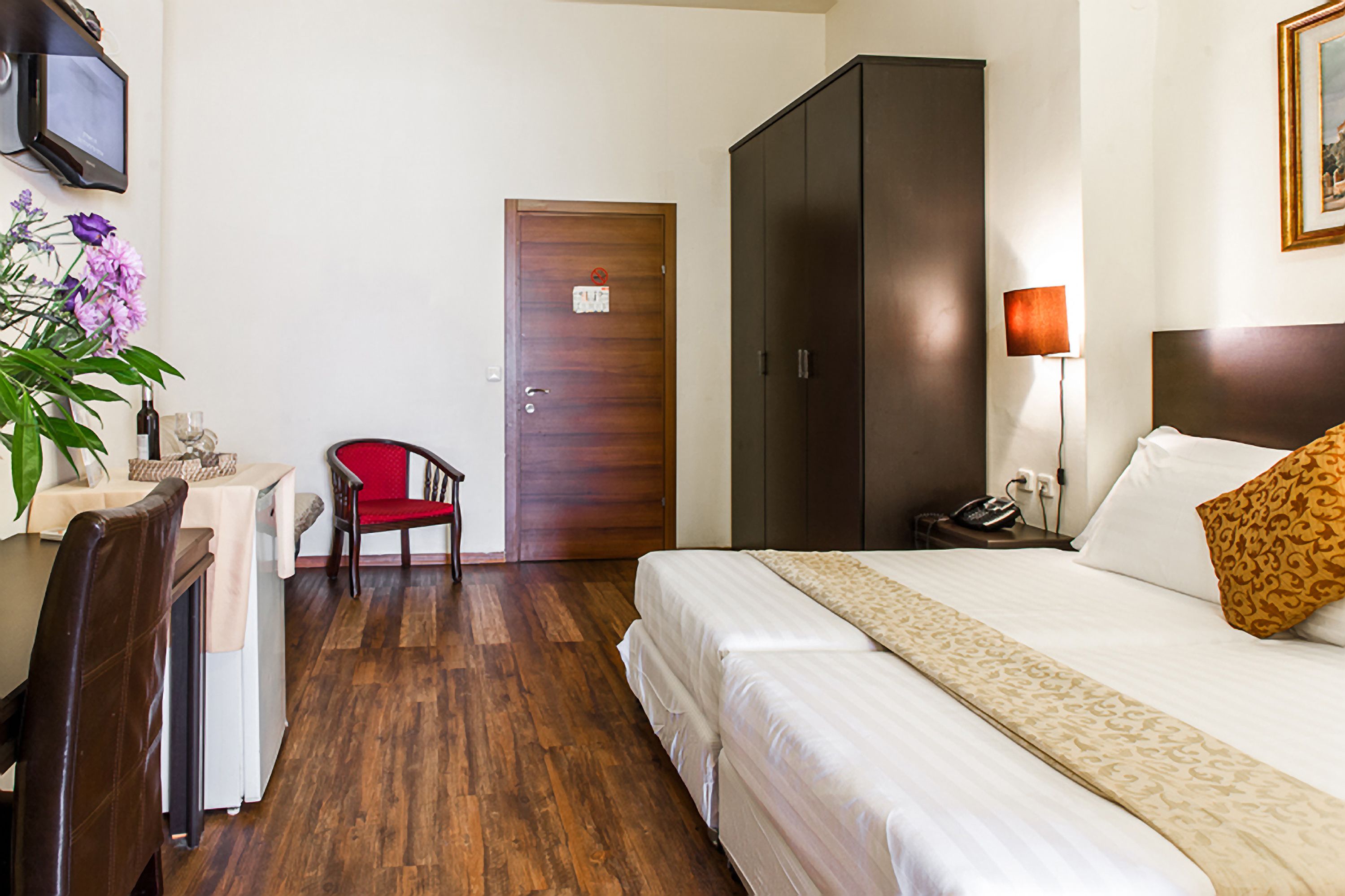 Una habitación de hotel con cama, escritorio con silla, armario, mesita de noche con lámpara y televisor en la pared.