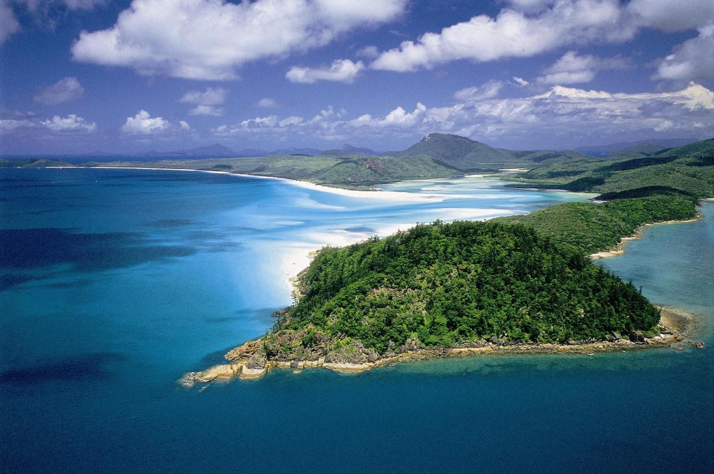 An aerial view of a coastal landscape with a beach, clear blue waters, and a forested island.