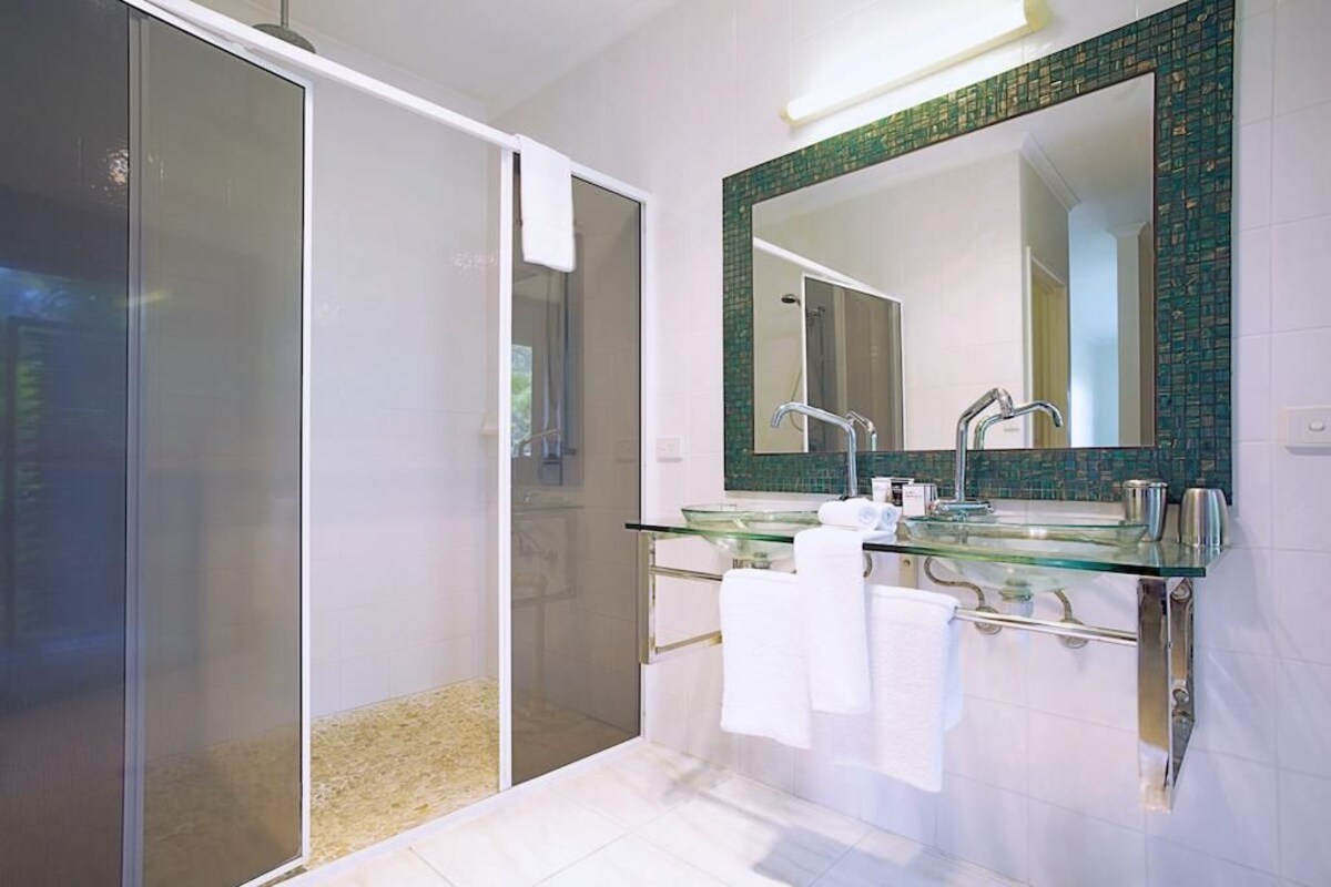A modern bathroom with a glass shower enclosure, a glass sink, and a large mirror with a green mosaic border.