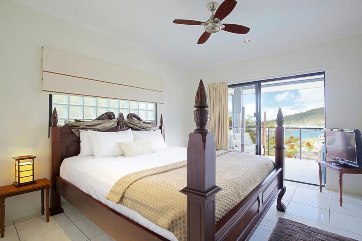 A bedroom with a large bed, a ceiling fan, a TV, and a balcony with a view.