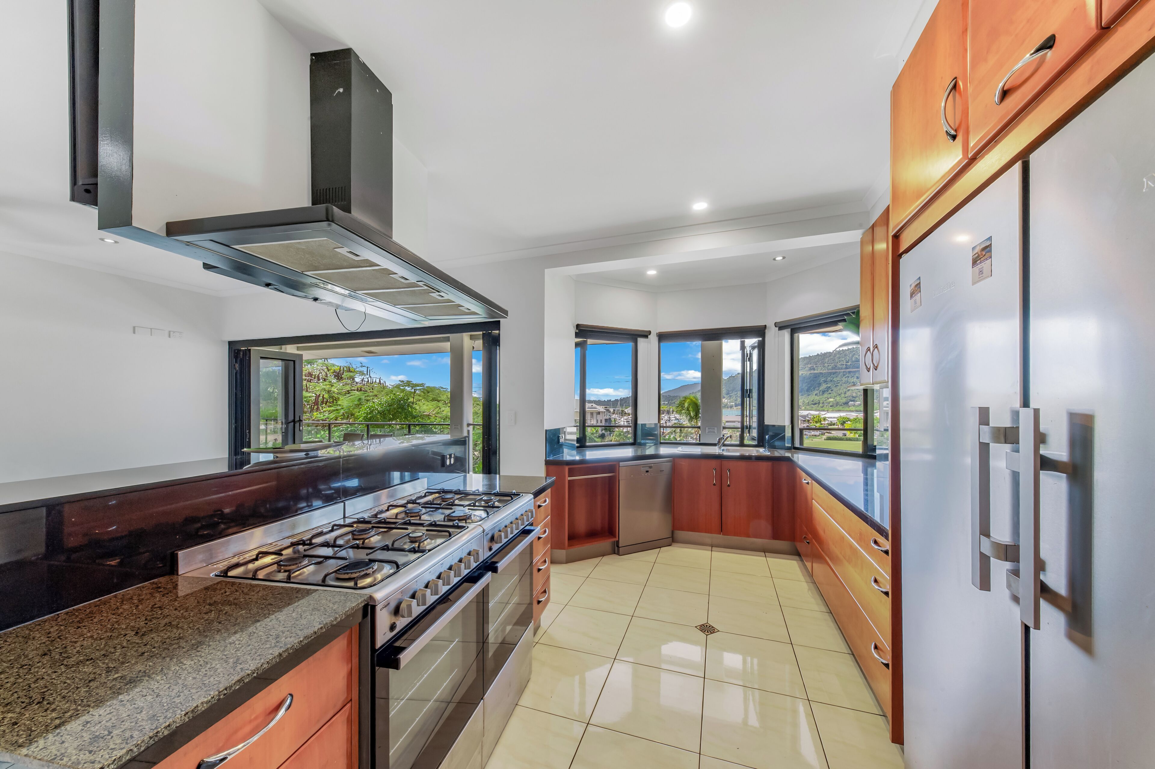 A modern kitchen with a gas stove, oven, stainless steel appliances, wooden cabinets, and large windows offering a view of greenery.