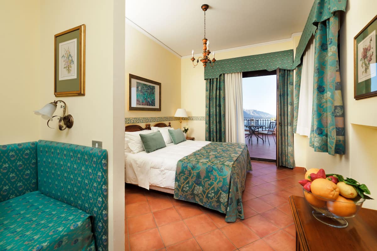 A hotel room with a bed, a sofa, a balcony with a table and chairs, and a fruit bowl.