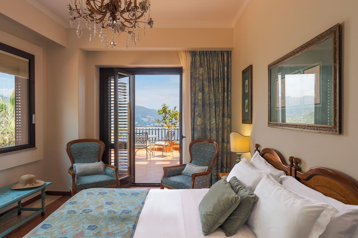 A hotel room with a bed, two armchairs, a chandelier, a balcony with a view, and a large mirror.