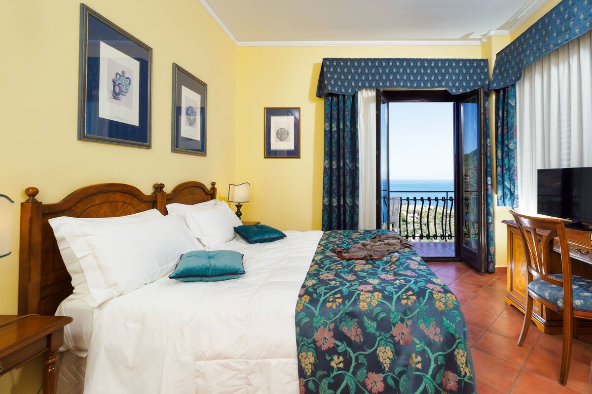 A hotel room with a bed, a desk with a chair, a television, and a balcony with a view of the sea.