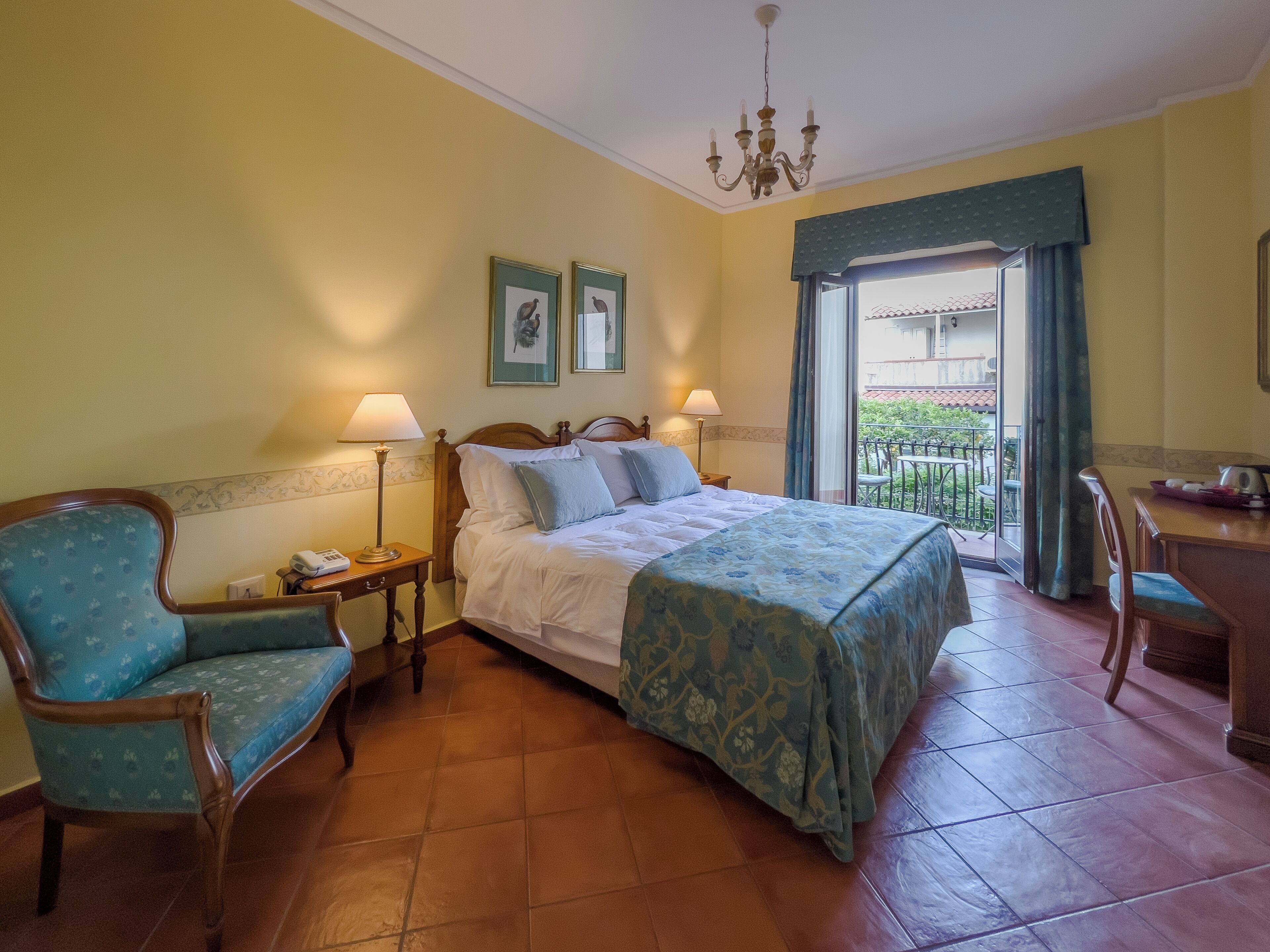 A hotel room with a bed, two bedside lamps, a chair, a small table, and a balcony with a view.