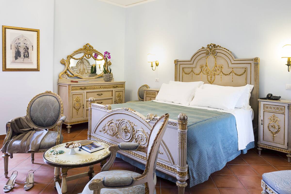 A classic, ornate bedroom with a large bed, a vanity with a mirror, and a small round table with a book and slippers.