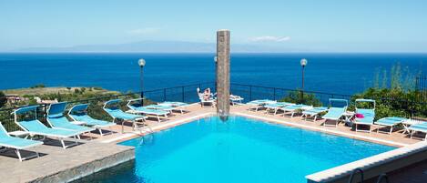 A pool with a clear view of the ocean, surrounded by lounge chairs and a stone pillar.