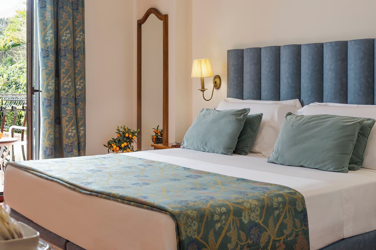 A hotel room with a large bed, a nightstand with a lamp, a mirror, and a view of the outdoors.