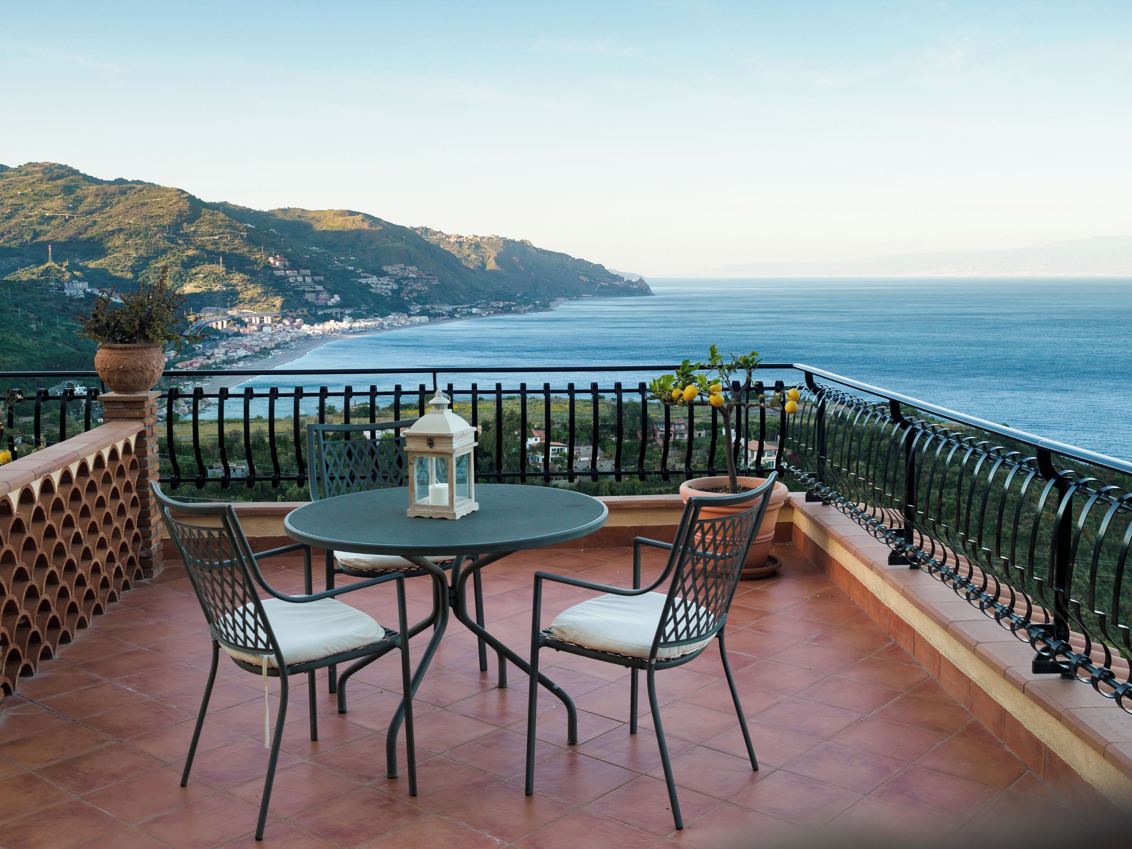 A terrace with a round table and two chairs, overlooking a coastal view with a mountain in the background.